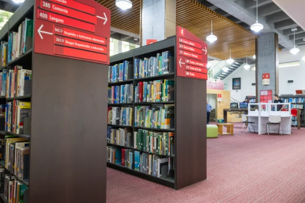Estanterías con libros organizados en una biblioteca moderna, con señalización roja que indica categorías y direcciones, y mesas de estudio al fondo en un espacio iluminado.