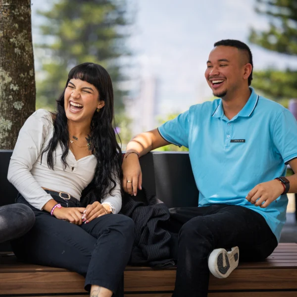 Dos personas están sentadas en una banca al aire libre, conversando en un espacio con árboles y edificios visibles en el fondo.
