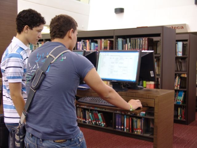 Dos personas están de pie frente a un computador en una biblioteca, consultando información en la pantalla con estanterías llenas de libros al fondo.