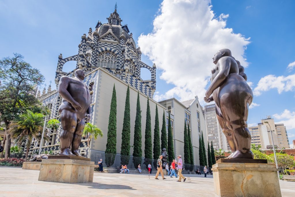 Plaza pública con esculturas de figuras humanas robustas en primer plano y al fondo el edificio histórico del Palacio de la Cultura Rafael Uribe Uribe, de arquitectura gótica, rodeado de árboles y cielo despejado.
