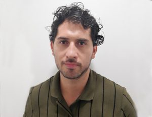 Persona con cabello oscuro y rizado, vestida con una camiseta verde con líneas verticales negras, frente a un fondo blanco uniforme.