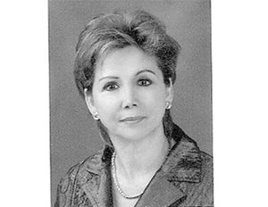 Persona con cabello corto y peinado hacia atrás, usando chaqueta formal con textura satinada y una cadena sencilla en el cuello, sobre un fondo gris uniforme.