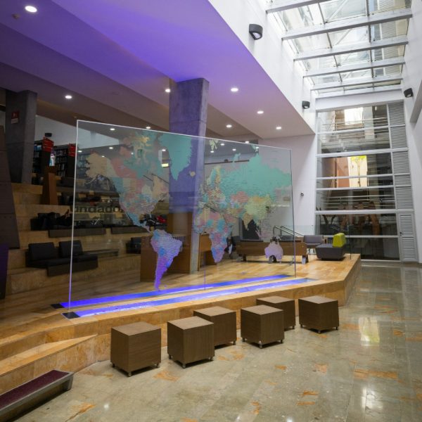 Vestíbulo amplio y moderno con techo de vidrio, iluminación natural y artificial. En el centro hay un panel transparente con un mapa mundial de colores, rodeado de asientos cúbicos. Al fondo se observan escaleras y áreas de descanso en niveles superiores.