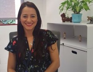 Persona sentada en una silla dentro de una oficina, usando una blusa oscura con estampado floral. Al fondo hay un mueble blanco con una planta en maceta azul y pequeñas figuras decorativas, junto a una ventana que deja entrar luz natural.