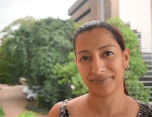 Persona en primer plano con cabello recogido, usando una prenda de tiras delgadas. Al fondo se observa vegetación frondosa y parte de un edificio moderno con paredes de ladrillo y ventanas, en un entorno exterior iluminado por luz natural.