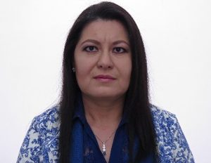 Persona con cabello oscuro y liso viste blusa blanca con estampado azul, frente a un fondo blanco.