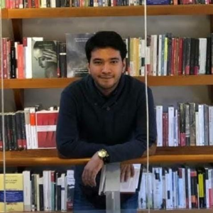 Persona de pie frente a una estantería llena de libros, sosteniendo un libro abierto sobre un mostrador.