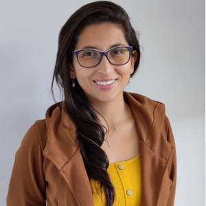 Persona con cabello largo y suelto viste camiseta amarilla y chaqueta marrón, de pie frente a un fondo gris claro.