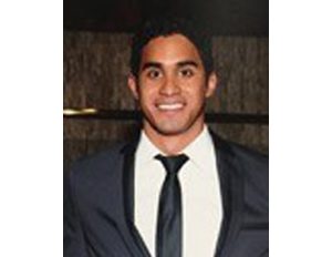 Persona vestida con traje formal oscuro, camisa blanca y corbata negra, frente a un fondo interior.