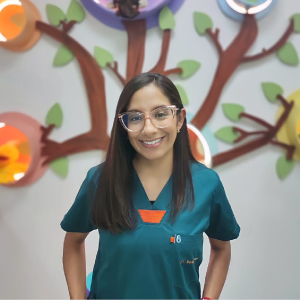 Persona con uniforme médico color verde oscuro está de pie frente a una pared decorada con figuras de árbol y círculos coloridos.