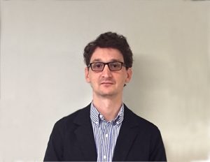 Persona vestida con camisa de rayas y saco oscuro, de pie frente a una pared clara.