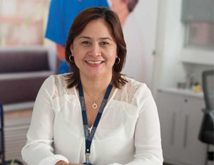 Persona con blusa blanca y gafete azul está sentada en un entorno interior, sobre una mesa clara, con un fondo que incluye una imagen difusa y mobiliario de oficina.