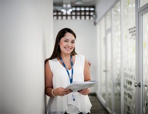 Persona con blusa blanca sin mangas y gafete azul sostiene documentos mientras está de pie en un pasillo interior con paredes blancas y puertas de vidrio.