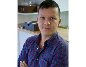 Persona con camisa de cuadros en tonos azul y rojo está sentada con los brazos cruzados, en un entorno interior donde se observa una caja y un recipiente etiquetado con la palabra “TE” sobre una repisa.