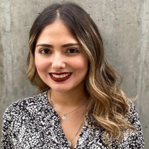 Persona con cabello largo y ondulado, parcialmente aclarado en las puntas, lleva una blusa estampada en blanco y negro y un collar sencillo, frente a un fondo gris liso.