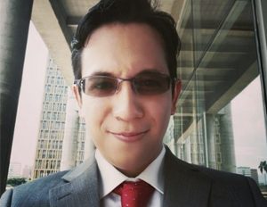 Persona con traje formal gris, camisa blanca y corbata roja, en un entorno urbano con columnas y edificios modernos al fondo.