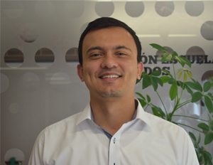 Persona con camisa blanca de botones, de pie en un entorno interior con pared decorada con círculos y una planta verde al fondo.