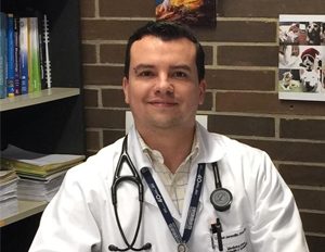 Persona con bata blanca de laboratorio, estetoscopio alrededor del cuello y gafete colgado, sentada frente a una pared de ladrillos con estantería de libros y documentos a un lado.