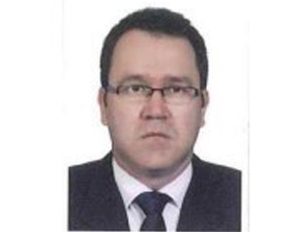 Persona vestida con traje formal oscuro, camisa blanca y corbata negra, frente a un fondo blanco.