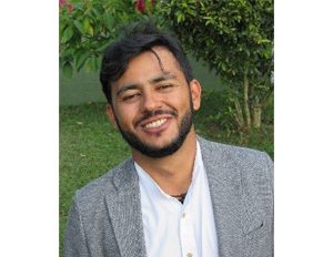 Persona vestida con camisa blanca y saco gris, de pie en un espacio exterior con césped y plantas en el fondo.