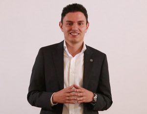 Persona vestida con traje formal oscuro y camisa blanca, de pie con las manos entrelazadas al frente, sobre fondo claro.