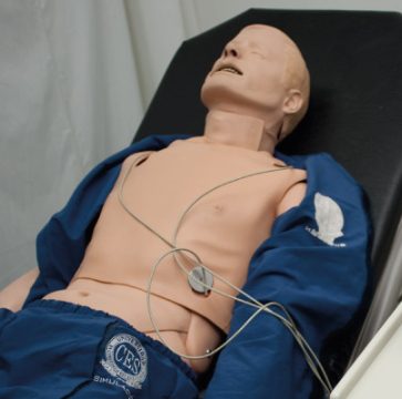 Simulador de cuerpo humano acostado en una camilla, con electrodos conectados al torso y vestido parcialmente con ropa azul, utilizado para prácticas médicas.