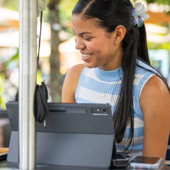 Persona sentada frente a una mesa usando una tableta con teclado. Al fondo se observan árboles y sombrillas en un espacio exterior.