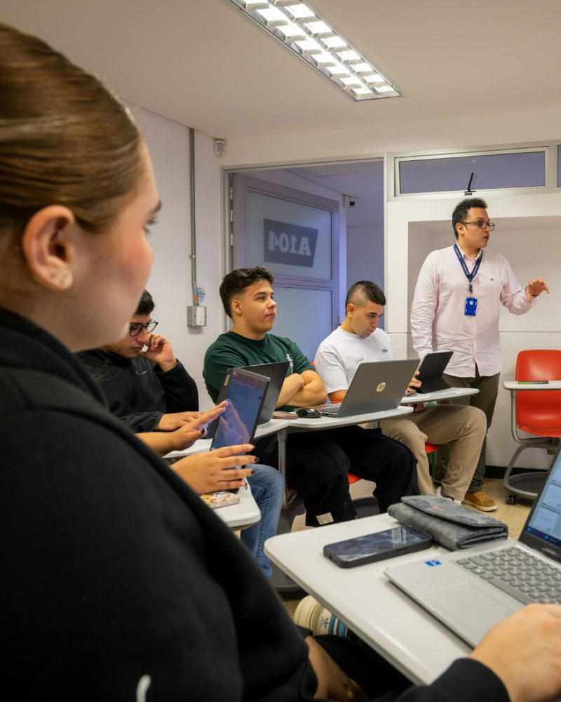 Persona de pie frente a un grupo en un aula, hablando mientras varias personas están sentadas con computadores portátiles y dispositivos sobre escritorios.