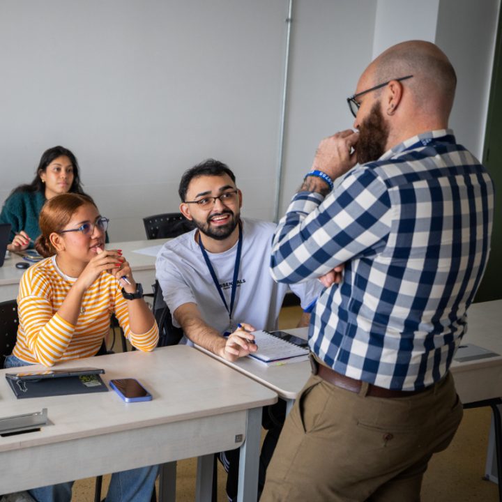 Persona de pie frente a estudiantes en un aula, conversando mientras los estudiantes toman notas en sus escritorios.