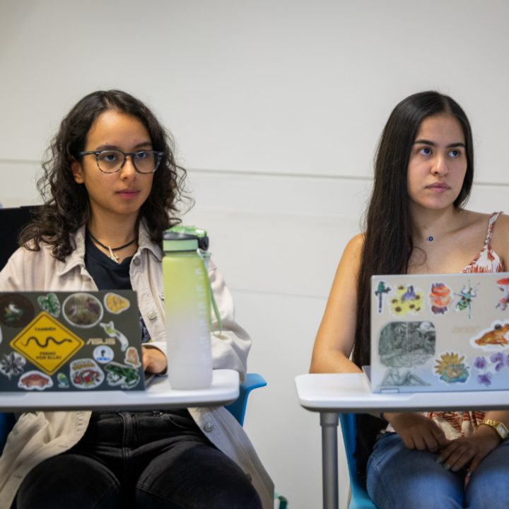 Dos personas están sentadas en escritorios con portátiles abierto. El fondo es una pared blanca lisa, en un entorno educativo.