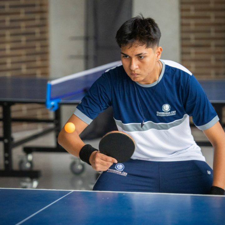 Persona jugando tenis de mesa, sosteniendo una raqueta y golpeando una pelota amarilla sobre una mesa azul. Al fondo se observa otra mesa de tenis de mesa y una pared de ladrillo.
