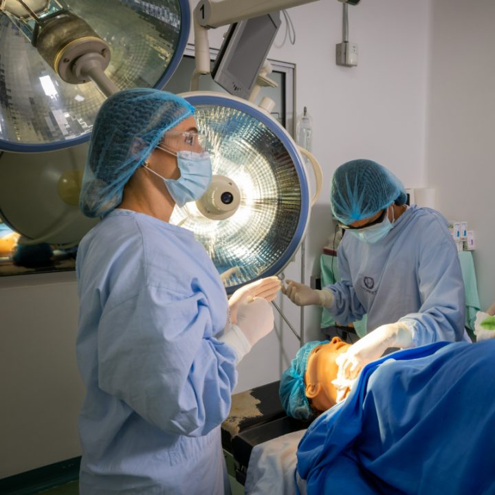 Dos profesionales de la salud realizan un procedimiento médico en un quirófano, usando batas, gorros y mascarillas. Hay lámparas quirúrgicas encendidas sobre la mesa donde se encuentra el paciente.