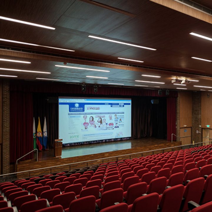 Auditorio con filas de sillas rojas frente a un escenario con cortinas rojas. En el escenario hay un podio y varias banderas, y se proyecta en una gran pantalla una página web con información educativa.