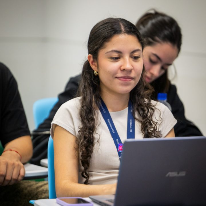 Persona sentada en un aula frente a un computador portátil, con cuadernos y bolígrafo sobre el escritorio. Al fondo se observan otras personas en un ambiente de clase.