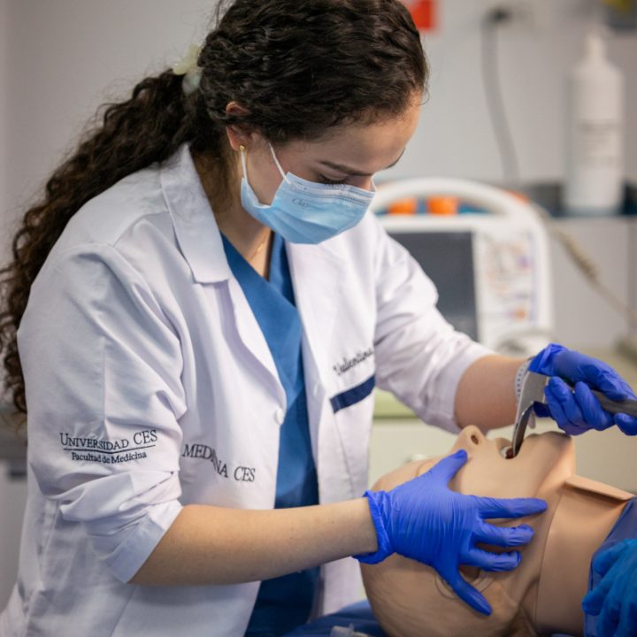Persona con bata de laboratorio y guantes realizando un procedimiento médico en un simulador clínico.