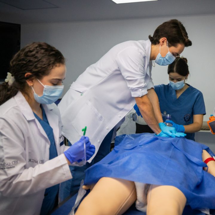 Tres personas con batas y mascarillas realizan un procedimiento médico sobre un simulador cubierto con una sábana azul en un entorno clínico.
