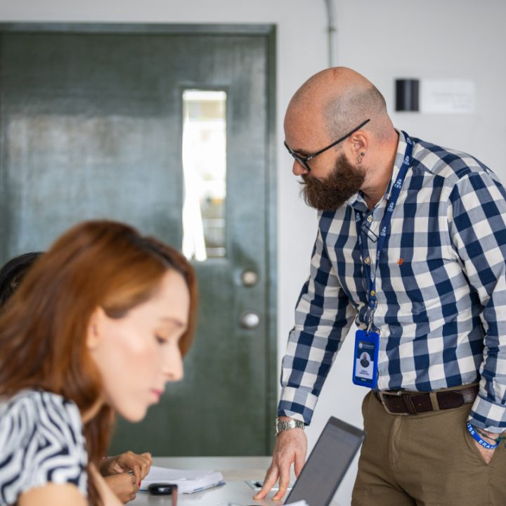 Persona de pie junto a una mesa en un aula, usando camisa de cuadros y portando un gafete; sobre la mesa hay un computador portátil y documentos, y al fondo se observa una puerta.