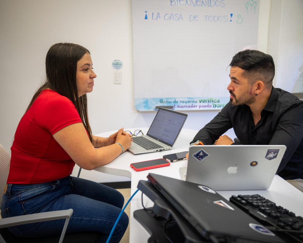 Dos personas están sentadas frente a frente en una mesa blanca, trabajando con computadores portátiles en una sala con tablero blanco al fondo.