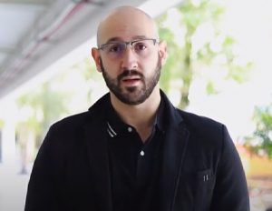 Persona con camisa negra y chaqueta oscura está de pie en un pasillo iluminado, con baranda y vegetación visible al fondo.