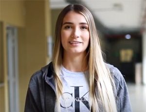 Persona con cabello largo y liso lleva puesta una camiseta blanca con el logo “CK” y una chaqueta gris, ubicada en un pasillo interior iluminado.