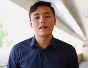 Persona con camisa azul está de pie en un pasillo iluminado, con paredes claras y una baranda al fondo.