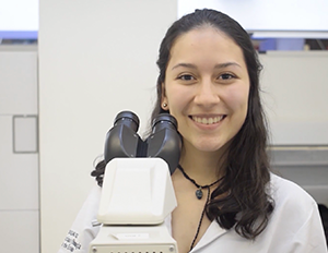 Persona con bata blanca está en un laboratorio frente a un microscopio, con fondo que incluye mobiliario y equipo científico.