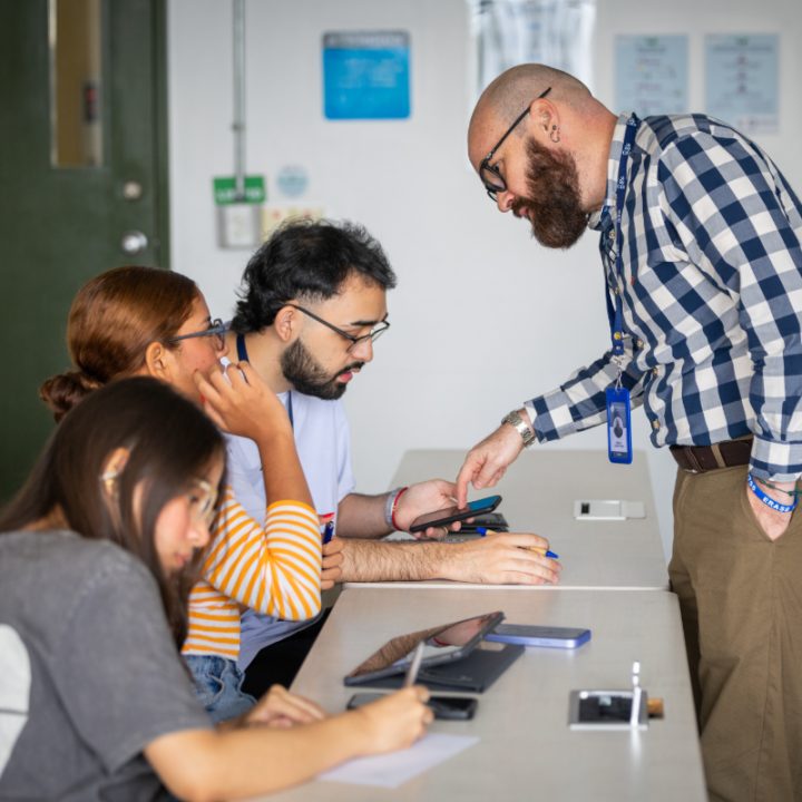 Persona de pie junto a un escritorio muestra algo en un dispositivo móvil a tres personas sentadas, que tienen cuadernos y bolígrafos sobre la mesa en un aula.
