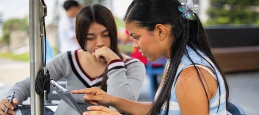 Dos personas están sentadas en un espacio al aire libre, interactuando con una tableta mientras conversan. Sobre la mesa hay cuadernos y bolígrafos.