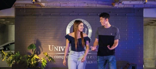 Dos personas caminan y conversan frente a una pared con el logo y nombre de la Universidad CES, en un espacio interior iluminado con luces moradas y plantas decorativas.
