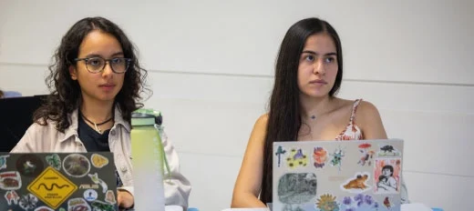 Dos personas están sentadas frente a computadores portátiles decorados con múltiples adhesivos coloridos, en un aula.