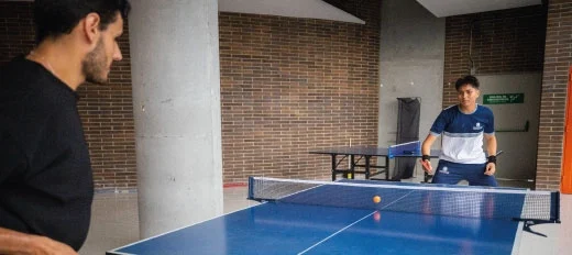 Dos personas juegan tenis de mesa en un espacio interior con paredes de ladrillo y columnas grises. La mesa es azul y la pelota está en movimiento sobre la red.