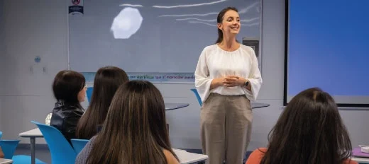 Persona de pie frente a un grupo en un aula, junto a una pantalla y un tablero, parece estar dando una presentación. Al fondo se observan sillas azules y mesas organizadas en el espacio educativo.