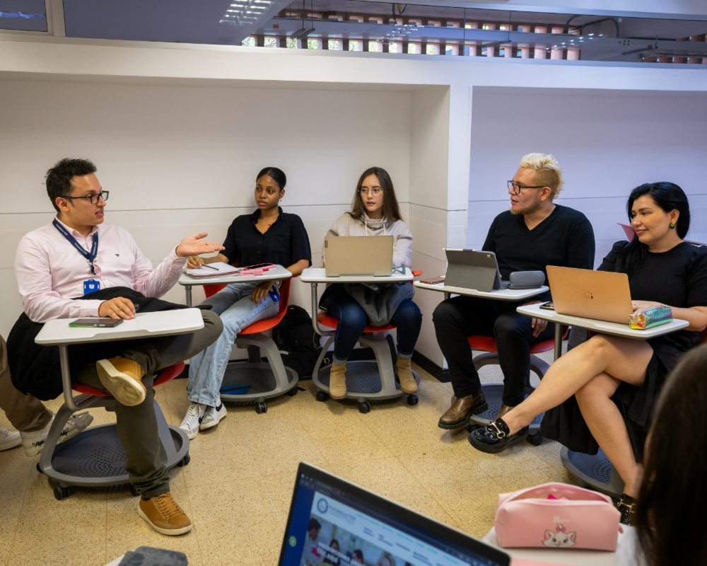 Grupo de personas sentadas en un aula en disposición circular, cada una con un computador portátil sobre escritorios individuales, mientras una persona dialoga con el resto del grupo.