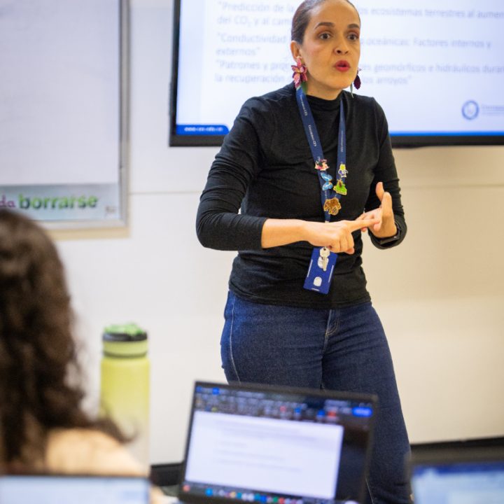 Persona de pie frente a una pantalla con texto proyectado, realizando una presentación en un aula mientras otras personas trabajan con portátiles.